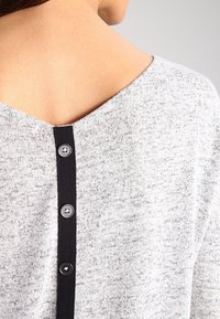 Szary dzianinowy top z dekoltem w serek, ozdobiony ciemnym, teksturowanym paskiem i czterema czarnymi guzikami z tyłu. Miękka, lekko melanżowa faktura.