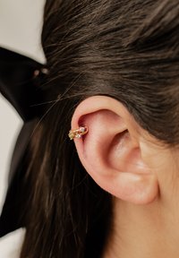 Gold Ohrring-Cuff mit gebogenem Design und kleinen klaren Edelsteinen. Am Ohr angebracht, im Kontrast zu braunem Haar. Glatte Textur.