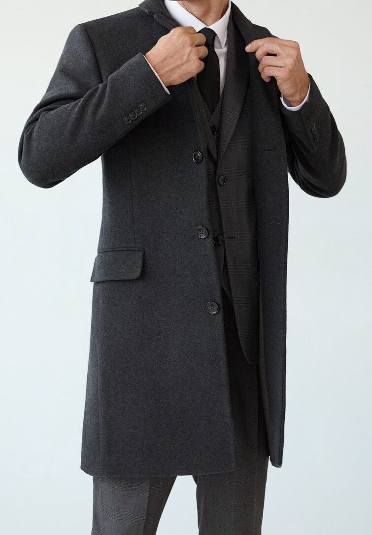 Lang, donkergrijze wollen jas met drie knopen, notch-kraag en zijzakken, gedragen over een zwart vest en stropdas, met een gladde textuur.