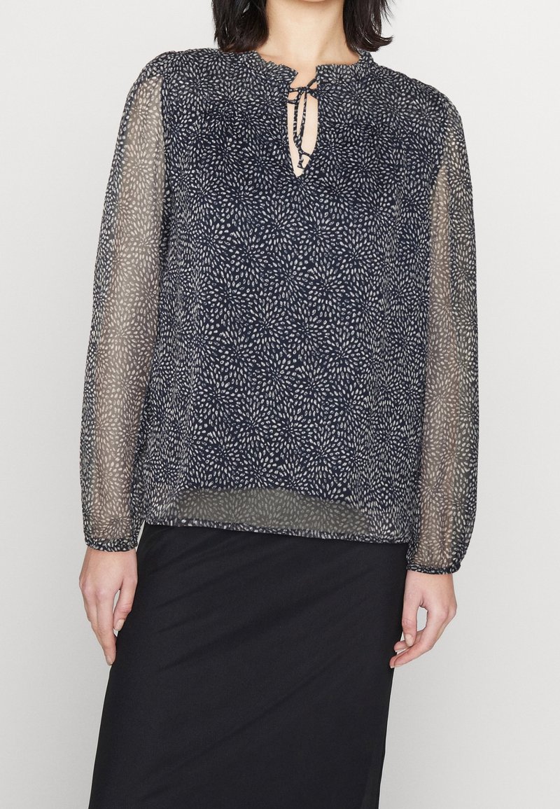 Blusa trasparente blu navy con un motivo floreale, scollatura a goccia e maniche lunghe raccolte ai polsi. Il tessuto è leggero e arioso.
