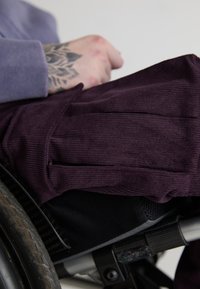 Pantaloni din catifea purpurie cu pliuri frontale, purtați de o persoană într-un scaun cu rotile. Materialul negru al scaunului cu rotile și anvelopa texturată din cauciuc sunt vizibile.