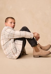 Beige ternet jakke med grå og brune accenter, sorte bukser og beige ruskindsstøvler med elastiske sidepaneler. Ingen bemærkelsesværdige mønstre.