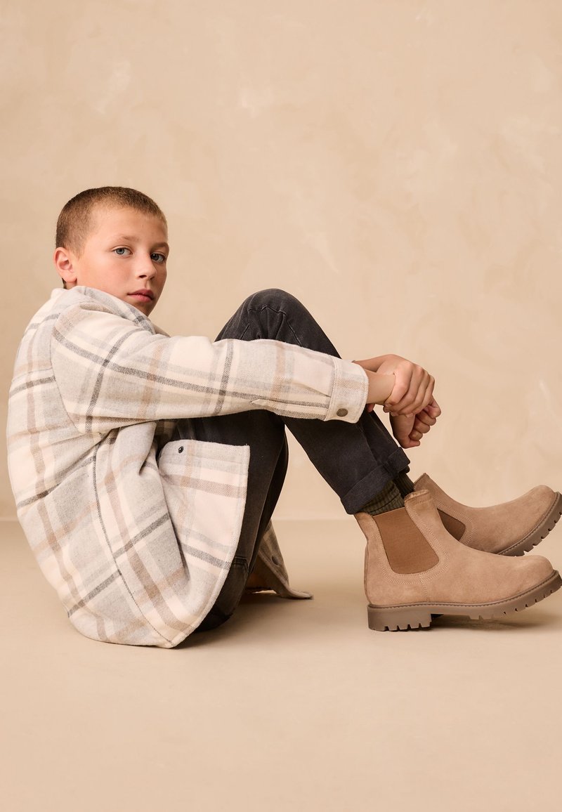 Beige karierter Blazer mit grauen und braunen Akzenten, schwarze Hose und beige Wildleder-Stiefeletten mit elastischen seitlichen Einsätzen. Keine auffälligen Muster.