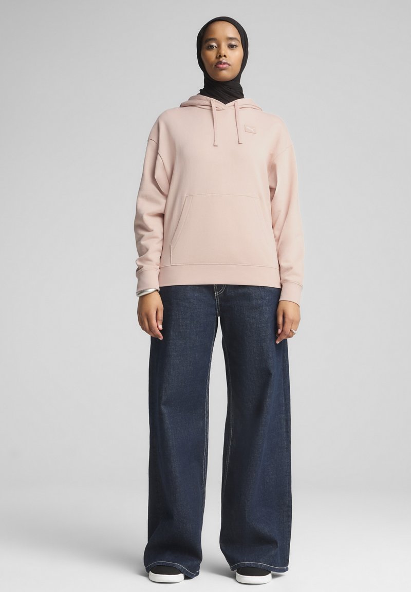 Sudadera con capucha de color rosa claro con un bolsillo tipo canguro. Combinada con jeans de pierna ancha de color azul oscuro. El modelo se encuentra tranquilo sobre un fondo gris sencillo.