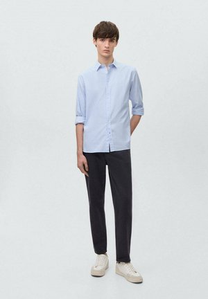 Hellblau gestreiftes Hemd mit Button-Down-Kragen, hochgekrempelten Ärmeln, kombiniert mit dunklen Hosen und weißen Sneakern vor einem neutralen Hintergrund.