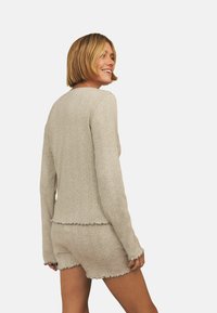 Kvinna med kort blont hår ler, iförd en beige ribbad långärmad topp och matchande shorts med vågiga kanter, stående mot en vit bakgrund.