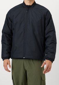 Giubbotto imbottito nero con colletto alto, zip frontale completa e polsini elastici. Indossato sopra pantaloni cargo oliva. Tessuto liscio e leggero.