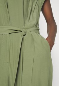 Mono de tela verde con cintura anudada, mangas cortas y bolsillos laterales. La textura es suave con detalles de pliegues sutiles.