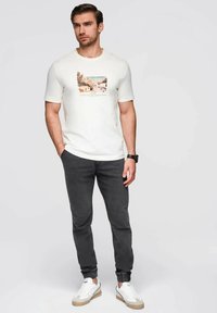 Camiseta de manga corta de hombre en color crema con un gráfico de paisaje impreso, combinada con joggers oscuros ajustados y zapatillas blancas con acentos en beige.