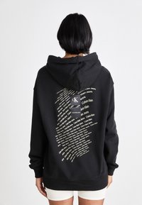 Svart huvtröja med texten "Calvin Klein" i upprepat tryck på baksidan, med avslappnad passform och ribbade ärmslut och nederkant.