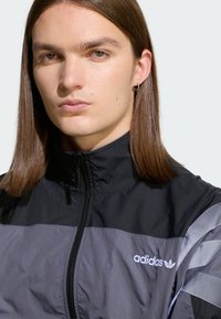 Čierna a sivá bunda na zips s vysokým golierom, vybavená kontrastným panelovým dizajnom a logom Adidas na hrudi. Hladký, ľahký materiál.