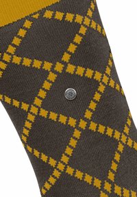 Brązowa skarpetka z musztardowo-żółtym wzorem w kratkę oraz małym srebrnym okrągłym emblematem z napisem i wzorem krzyża.