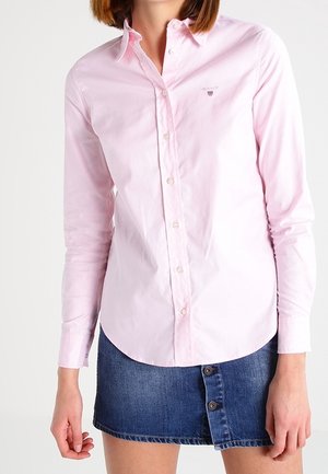 Woman wearing a light pink button-up shirt and a blue denim mini skirt with front buttons, standing against a plain background.