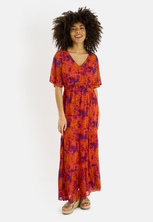 Femme aux cheveux bouclés portant une longue robe maxi fluide à motif orange et violet avec un décolleté en V, et des sandales à plateforme beige, souriante.