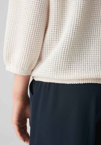 Weißes, strukturiertes Strickoberteil mit Dreiviertelärmel, elastischen Bündchen und geradem Saum, kombiniert mit dunkelblauen Hosen.