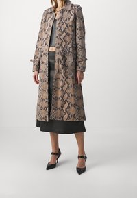 Trench-coat long à motif peau de serpent en tons beige et marron, avec col, boutons sur le devant et poignets ceinturés. Associé à des talons noirs.