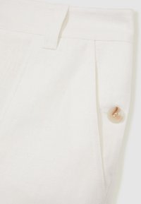 Close-up of white linen pants waistband with belt loop, side pleat, side pocket, and beige button detail.