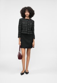 Sort/hvid ternet cropped jakke med guldknapper, parret med en sort nederdel. Modellen holder en bourgogne håndtaske og har sorte flade sko på.