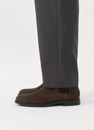 Bruine suède Chelsea-laarzen gedragen met donkergrijze broeken tegen een effen witte achtergrond.