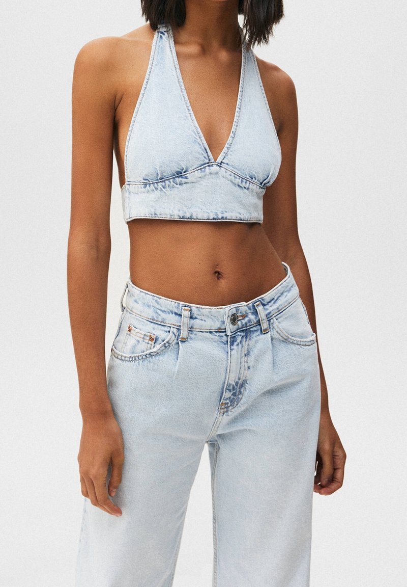 Femme portant un crop top halter en jean bleu clair et un jean taille haute assorti, debout devant un fond uni.