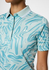 Jasnoniebieska koszulka polo z falistym wzorem w ciemniejszym odcieniu niebieskiego, z dwuguzikowym dekoltem i prążkowanym kołnierzem ozdobionym białymi paskami.