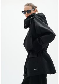 Femme portant un manteau à capuche noir et des lunettes de soleil noires angulaires, posant les mains dans les poches devant un fond clair uni.
