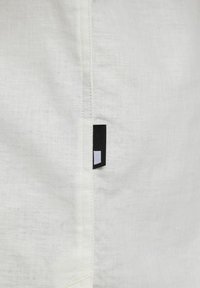 Tessuto di lino bianco con texture visibile, caratterizzato da un'etichetta nera con un piccolo quadrato bianco sul bordo. Design semplice e pulito.