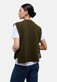 Gilet sans manches vert olive en tricot avec des bords côtelés, superposé sur un t-shirt blanc, associé à un jean bleu foncé, vu de dos.