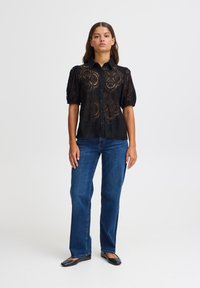 Camicia nera in pizzo con maniche a sbuffo, caratterizzata da intricate fantasie circolari, abbinata a jeans a gamba dritta blu e ballerine nere.