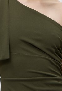 Κοντινή λήψη ενός ελιάς πράσινου φόρεματος με έναν ώμο, με ριγωτό ύφασμα και ασύμμετρο ντεκολτέ στο πάνω μέρος του κορμού.