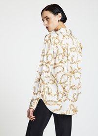 Weiße Bluse mit einem goldenen Kettenmuster, langen Ärmeln und einem Kragen. Verfügt über eine lockere Passform und einen abgerundeten Saum aus glattem Stoff.