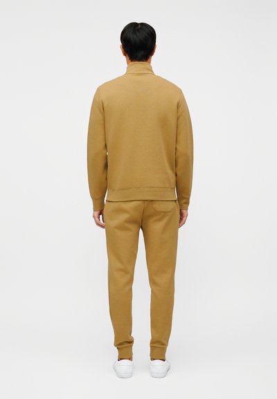 Άνδρας με ταίριαστο μπεζ φούτερ και παντελόνι φόρμας, φορώντας λευκά αθλητικά παπούτσια, στέκεται με την πλάτη προς τα εμπρός σε ένα απλό λευκό φόντο.