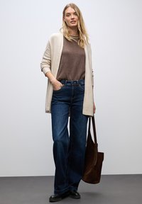 Cardigan beige sopra un maglione marrone, abbinato a jeans a gamba larga blu. L'outfit include una borsa a tracolla marrone e calzature nere.
