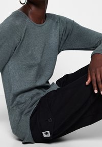 Harmaa pitkähihainen paita, joka on valmistettu kevyest ä kankaasta. Mustat housut, joissa on nappisulku ja logopatchi taskussa. Yksinkertainen, rento istuvuus.