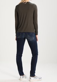 Olivový top s dlouhým rukávem v kombinaci s úzkými tmavě modrými džíny a stříbrnými teniskami. Tento outfit má jednoduchý design bez viditelných vzorů.