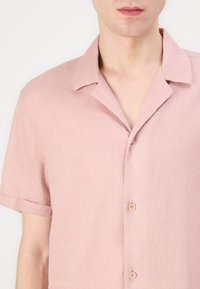 Camicia corta a maniche corte di colore rosa chiaro, realizzata in tessuto texturizzato, con colletto a causa e due bottoni sul davanti.