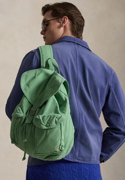 Άνδρας που φοράει γυαλιά ηλίου, μπλε μπουφάν και μπλε παντελόνι μεταφέρει πράσινο καμβά σακίδιο στον ώμο του, με ουδέτερο φόντο.