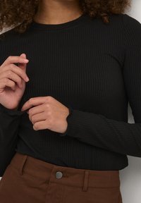 Czarny prążkowany top z długim rękawem o okrągłym dekolcie, z dopasowanymi mankietami i teksturowanym wzorem. Brązowe spodnie z metalowym zapięciem.