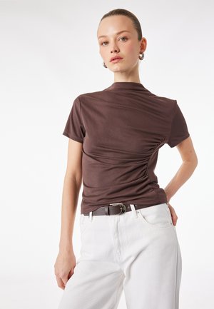 Frau mit zurückgekämmtem Haar trägt ein eng anliegendes braunes Kurzarmoberteil mit seitlicher Raffung und weiße hochtaillierte Hosen mit einem dunklen Gürtel.