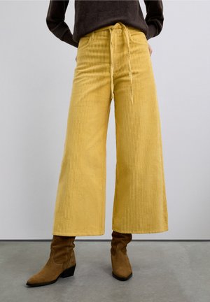 Pantalones anchos de pana amarilla con cintura ajustable con cordón y textura acanalada vertical, combinados con botines marrones de ante.