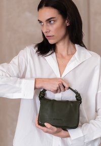 Borsa in pelle verde con manico annodato, caratterizzata da una finitura morbida e testurizzata e da una forma rettangolare. La modella la tiene vicino a una camicia bianca.