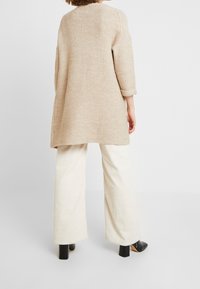 Tunique en tricot beige texturé avec des manches trois-quarts, associée à un pantalon large en velours côtelé crème, porté avec des chaussures à talons noires.