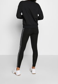 Czarna bluza z okrągłym dekoltem i ściągaczem na dole, zestawiona z czarnymi legginsami z białymi paskami po bokach i akcentami logo. Białe buty sportowe.