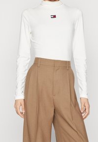 White long-sleeve turtleneck top with a small black, red, and white logo patch. Paired with high-waisted, light brown pleated trousers.