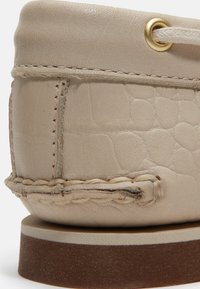 Τακούνι παπούτσι από μπεζ δέρμα με υφή, ορατή ραφή, πλεγμένη λεπτομέρεια, κορδόνι σχοινί μέσα από μπρούτζινο οφθαλμό και στρωμένο καφέ πέλμα.