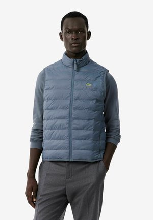 Man draagt een lichtblauw doorgestikt vest over een blauw overhemd met lange mouwen en een grijze broek, staand met één hand in de zak.