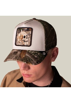 Joven lleva una gorra de béisbol beige y de camuflaje con un parche de calavera con la etiqueta "CANCELLED" en el panel frontal.