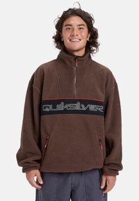 Sudadera de forro polar marrón con cuello alto, cierre de media cremallera, detalles rojos y una banda negra con el logo de "Quiksilver". Incluye bolsillos laterales.
