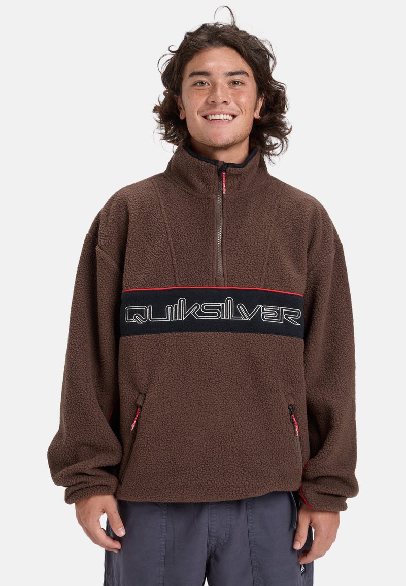Sudadera de forro polar marrón con cuello alto, cierre de media cremallera, detalles rojos y una banda negra con el logo de "Quiksilver". Incluye bolsillos laterales.