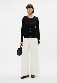 Cardigan nero con bottoni dorati, indossato sopra pantaloni bianchi a gamba larga. Gli accessori includono una piccola borsa intrecciata nera e stivali neri con tacco.
